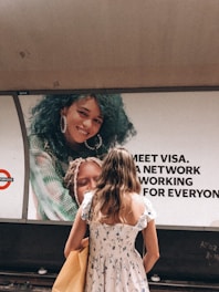 A woman in a floral dress stands in front of a large advertisement featuring a smiling person with curly blue hair. There are bold letters next to the image, part of an advertisement likely related to Visa. The setting appears to be a subway or train station.