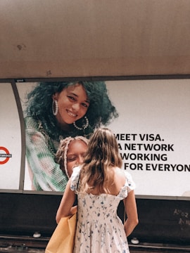 A woman in a floral dress stands in front of a large advertisement featuring a smiling person with curly blue hair. There are bold letters next to the image, part of an advertisement likely related to Visa. The setting appears to be a subway or train station.