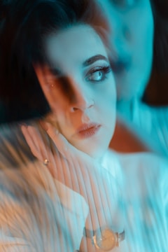 A portrait of a woman with a dreamy, ethereal quality achieved through soft focus and reflections. Her hand rests on her cheek, and she wears a wristwatch. The image has a blurred, artistic overlay with a prominent use of cool and warm tones.
