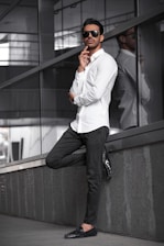 A casually dressed man leaning against a light-colored wall in a sunlit urban setting, embodying effortless style.