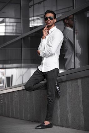 A casually dressed man leaning against a light-colored wall in a sunlit urban setting, embodying effortless style.