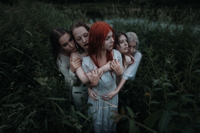 Five people stand closely together in a lush, overgrown field. They are embracing each other with somber expressions, creating an intimate and connected atmosphere. The greenery surrounding them is dense, adding a sense of enclosure and seclusion.