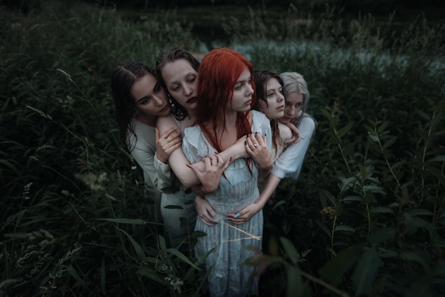 Five people stand closely together in a lush, overgrown field. They are embracing each other with somber expressions, creating an intimate and connected atmosphere. The greenery surrounding them is dense, adding a sense of enclosure and seclusion.