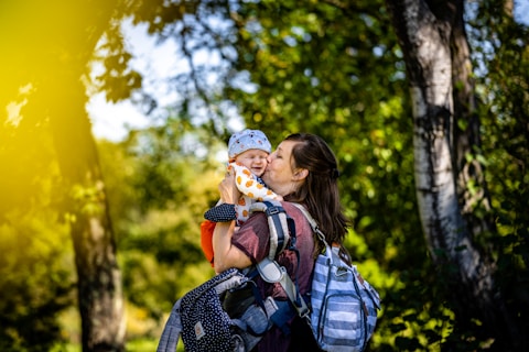 A woman is holding and kissing a baby in a bright and lush forest setting. The baby is wearing a patterned hat and an orange outfit. Both appear happy and content. They are surrounded by green foliage and trees, with sunlight filtering through, creating a warm glow.