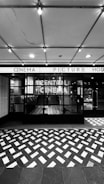 A black and white photograph of a cinema entrance featuring a large sign reading 'Central Cinema'. The façade is modern with large glass panes, and an awning with lit signage above displaying 'Cinema Picture House'. The floor outside is decorated with a geometric pattern of light and dark tiles. Visible inside are staircases and part of the interior.