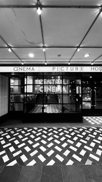 A black and white photograph of a cinema entrance featuring a large sign reading 'Central Cinema'. The façade is modern with large glass panes, and an awning with lit signage above displaying 'Cinema Picture House'. The floor outside is decorated with a geometric pattern of light and dark tiles. Visible inside are staircases and part of the interior.