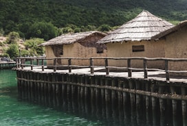 A series of rustic huts with thatched roofs are situated on a wooden platform above a body of greenish-blue water. The huts are constructed from natural materials, likely straw and mud, and are surrounded by lush green hills and dense foliage.