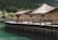 A series of rustic huts with thatched roofs are situated on a wooden platform above a body of greenish-blue water. The huts are constructed from natural materials, likely straw and mud, and are surrounded by lush green hills and dense foliage.