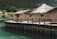 A series of rustic huts with thatched roofs are situated on a wooden platform above a body of greenish-blue water. The huts are constructed from natural materials, likely straw and mud, and are surrounded by lush green hills and dense foliage.