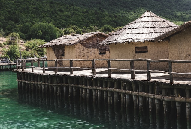 A series of rustic huts with thatched roofs are situated on a wooden platform above a body of greenish-blue water. The huts are constructed from natural materials, likely straw and mud, and are surrounded by lush green hills and dense foliage.