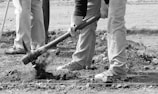 Several individuals are working together, focusing on digging or gardening. One person is using a pickaxe to break the ground, with soil and rocks visible. The scene conveys a sense of manual labor in an outdoor setting.