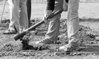 Several individuals are working together, focusing on digging or gardening. One person is using a pickaxe to break the ground, with soil and rocks visible. The scene conveys a sense of manual labor in an outdoor setting.