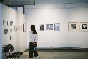 A woman stands in an art gallery observing framed photographs and artwork displayed on white walls. Various art pieces in different sizes are hung in a neat row. Empty chairs are positioned against the wall, suggesting additional seating or use for events.