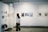 A woman stands in an art gallery observing framed photographs and artwork displayed on white walls. Various art pieces in different sizes are hung in a neat row. Empty chairs are positioned against the wall, suggesting additional seating or use for events.