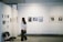 A woman stands in an art gallery observing framed photographs and artwork displayed on white walls. Various art pieces in different sizes are hung in a neat row. Empty chairs are positioned against the wall, suggesting additional seating or use for events.