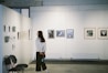 A woman stands in an art gallery observing framed photographs and artwork displayed on white walls. Various art pieces in different sizes are hung in a neat row. Empty chairs are positioned against the wall, suggesting additional seating or use for events.