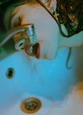A person is drinking water directly from a faucet, positioned over a sink. The image has a blue-green tint, and the lighting creates a moody, almost surreal effect. The person has wet hair, and a cross-shaped earring is visible.