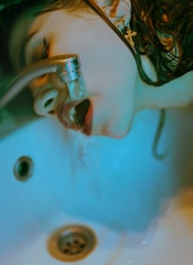 A person is drinking water directly from a faucet, positioned over a sink. The image has a blue-green tint, and the lighting creates a moody, almost surreal effect. The person has wet hair, and a cross-shaped earring is visible.