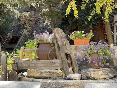 A rustic outdoor garden area features large stone planters filled with colorful flowers. The wooden structure blends with the natural elements, including surrounding trees and lush greenery. Steps lead up into the wooded area, offering a serene and inviting atmosphere.
