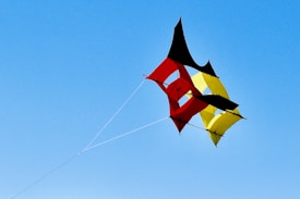 A colorful geometric kite with red, yellow, and black sections is flying against a clear blue sky. The kite has a unique design, composed of various interlocking shapes that create a dynamic structure, and is attached to a thin string.