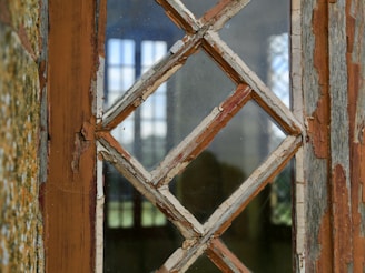 A close-up of an old, weathered window with peeling paint and a diamond-shaped wooden lattice frame. The glass panes are slightly dirty and reflect light from the outside.