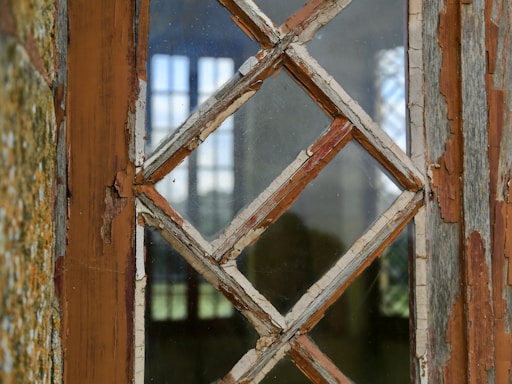A close-up of an old, weathered window with peeling paint and a diamond-shaped wooden lattice frame. The glass panes are slightly dirty and reflect light from the outside.
