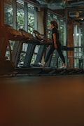 A person is walking on a treadmill in a dimly lit gym with large windows providing natural light. The individual is dressed in athletic wear and the indoor plants outside the windows add a touch of nature to the modern gym environment.