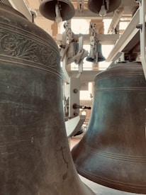 Large metal bells with decorative engravings are suspended in a bell tower. The bells are part of a mechanical assembly with levers and support structures visible. The sunlight filters through creating a warm ambiance.