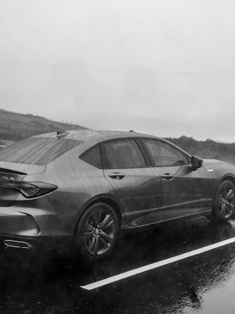 A sleek sedan is parked in a wet outdoor setting with rain visibly falling. The ground is reflective with water, and the scenery is somewhat obscured by fog or mist. The car's surface is shiny, indicating recent rainfall.