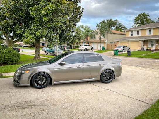 A sleek, silver sedan is parked on a driveway in a suburban neighborhood. The car has tinted windows and custom black rims. Surrounding the driveway are well-maintained lawns with green grass and trees. Multiple homes with two-car garages are visible, and other parked vehicles can be seen in the neighborhood.