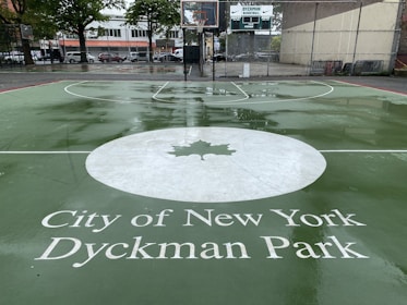 A wet and shiny outdoor basketball court with green paint and white lines featuring a design of a large white circle with a maple leaf in the center. The text 'City of New York Dyckman Park' is prominently displayed in the foreground. In the background, there is a basketball hoop, scoreboard, and urban buildings with some parked cars and trees.