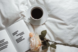 A cup of coffee sits on a white saucer atop a soft, white fabric surface. Next to it, an open book with poetic text lies flat, accompanied by a delicate, pale pink rose. The arrangement suggests a peaceful and reflective moment.