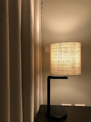 A modern table lamp with a fabric lampshade emits a warm glow next to beige curtains. The lamp stands on a dark wooden surface, and a light switch is visible on the wall behind.