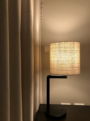 A modern table lamp with a fabric lampshade emits a warm glow next to beige curtains. The lamp stands on a dark wooden surface, and a light switch is visible on the wall behind.