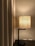 A modern table lamp with a fabric lampshade emits a warm glow next to beige curtains. The lamp stands on a dark wooden surface, and a light switch is visible on the wall behind.