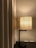 A modern table lamp with a fabric lampshade emits a warm glow next to beige curtains. The lamp stands on a dark wooden surface, and a light switch is visible on the wall behind.