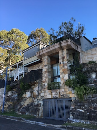 A modern house with a stone facade built on a rocky hillside. The stonework is complemented by metal railings and the structure features large glass windows. There's lush greenery with plants on the balcony and some trees in the background. The sky is clear and blue.