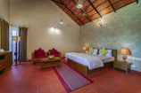 A cozy and well-lit bedroom with a double bed featuring white bedding and green accent pillows. The room includes wooden furniture such as bedside tables with lamps, a coffee table, and seating with red cushions. The floor is tiled with terracotta tiles, and the ceiling has a decorative pattern with an overhead fan. Natural light comes through sheer curtains next to heavy drapes, and a standing lamp provides additional illumination.