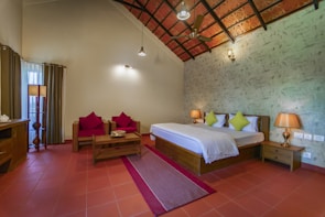 A cozy and well-lit bedroom with a double bed featuring white bedding and green accent pillows. The room includes wooden furniture such as bedside tables with lamps, a coffee table, and seating with red cushions. The floor is tiled with terracotta tiles, and the ceiling has a decorative pattern with an overhead fan. Natural light comes through sheer curtains next to heavy drapes, and a standing lamp provides additional illumination.