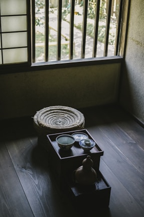 A traditional Japanese room featuring a wooden floor, low table with a ceramic bowl and small dish, a decorative gourd, and a woven round cushion. Sunlight filters through wooden slats of a window, casting shadows on the floor. The room exudes simplicity and tranquility.