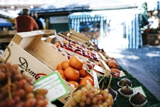 A vibrant display of fresh fruits arranged in wooden crates at an outdoor market. The foreground features oranges and various other fruits, with figs displayed in small paper cups nearby. In the background, people are visible, adding a sense of community and activity, while blue and white striped canopies provide shade.