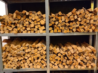 Several stacks of neatly chopped firewood are arranged on metal shelves. The wood pieces have a natural, rustic appearance with varying shades of brown and some visible bark. The shelves are organized in a neat and orderly fashion.