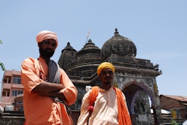 Two individuals dressed in traditional attire stand in front of an intricately carved, historic stone temple. The temple displays detailed architecture with prominent domes and decorative carvings. The individuals wear orange outfits with head coverings and carry small bags.