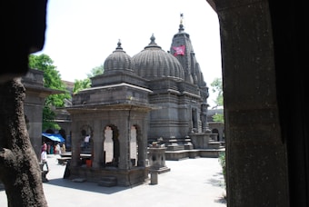 An ornate stone temple with intricate carvings and a distinct dome structure is set in an open courtyard. The architecture features detailed reliefs and ornamental towers. People are walking around the temple, adding to the serene and spiritual atmosphere. Trees with lush green foliage surround the structure, and sunlight casts shadows on the stone surfaces.