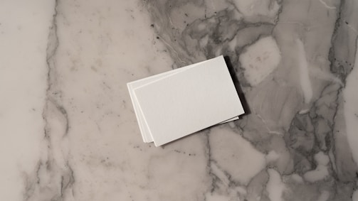 A stack of white business cards is placed on a grey and white marble surface. The surface displays a natural pattern with a mix of light and dark grey tones, creating a sleek and polished look.