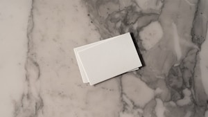 A stack of white business cards is placed on a grey and white marble surface. The surface displays a natural pattern with a mix of light and dark grey tones, creating a sleek and polished look.