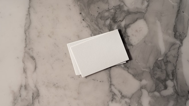 A stack of white business cards is placed on a grey and white marble surface. The surface displays a natural pattern with a mix of light and dark grey tones, creating a sleek and polished look.