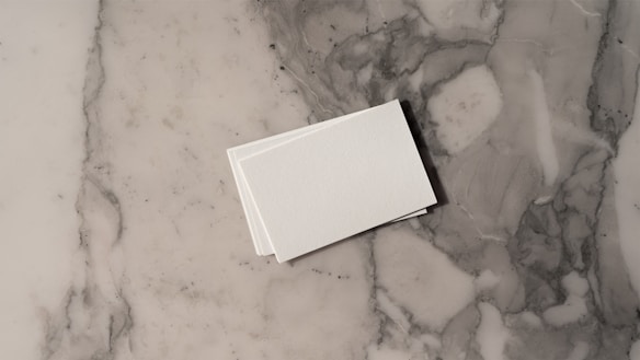 A stack of white business cards is placed on a grey and white marble surface. The surface displays a natural pattern with a mix of light and dark grey tones, creating a sleek and polished look.