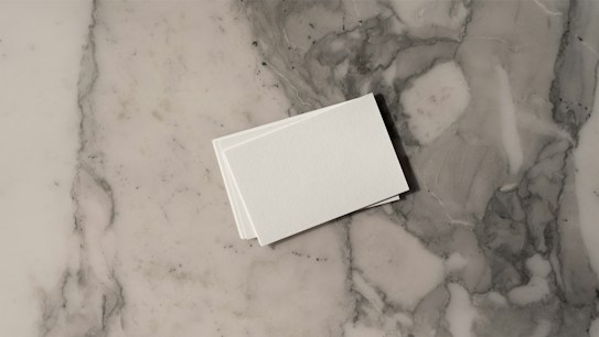 A stack of white business cards is placed on a grey and white marble surface. The surface displays a natural pattern with a mix of light and dark grey tones, creating a sleek and polished look.