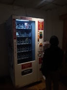 A dimly lit vending machine with various compartments, some containing products like snacks. A person stands in front of the machine, silhouetted in the darkness. The machine has instructions and pricing information displayed in different languages.